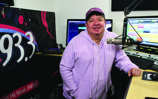 Man leaning on a stand up desk in front of a radio personality microphone and computers